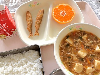 ピンクのトレーに給食が左から時計回りに牛乳、わかさぎ天ぷら（2こ）、半分にカットされたみかん、マーボー豆腐、ごはんが並んでいる写真