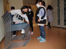 黒いマスクをした女の子が椅子を重ねて置く様子を白いマスクをした女の子が見ている写真