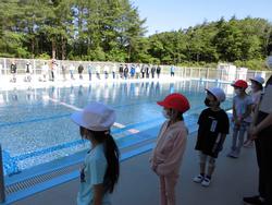 学校の屋外プールサイドで、紅白帽をかぶった子どもたちが先生と向かい合って整列している写真
