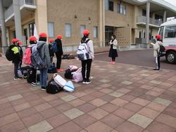 屋外で赤い帽子をかぶった子どもたちが荷物を足元に置いて並び、引率の先生らしき大人の話を聞いている様子の写真