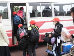 赤い帽子をかぶった子どもたちが大きな荷物を持って順番にバスへ乗り込んでいる写真