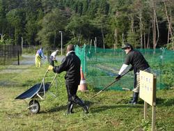 複数の人々が作業着や長靴を着用し、一輪車や鍬などの道具を使って作業を行っている様子の写真