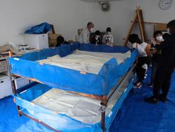 室内にブルーシートを敷き、その上に二段の大きな寝床が置かれ、周囲では子どもや大人が集まって観察や作業をしている写真