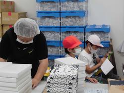 白い作業帽とマスクをつけた工場の人が箱詰め作業をしている横で、赤と白の帽子をかぶった子どもたちがノートにメモを取っている写真