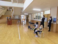 明るいフローリングの広いホールで、男性が三脚に設置されたタブレットに向かって話し、手前には代表の生徒2人が座り、その後ろで教員らしき男女2人が立って見守る始業式での写真