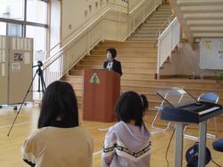 階段の下に設置された演台に立つ女性の話を聞いている生徒2名を後方から写した写真