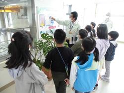 作業服を着た関係者が資料を指差し説明するのを子供達が真剣に聞いている様子の写真