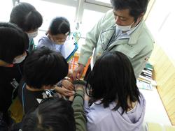 作業服を着た関係者の話を周りに集まった児童達が興味深く聞いている様子の写真