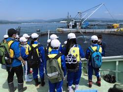 白いヘルメットとライフジャケットを身につけた子どもたちが船の上から大きな生け簀と作業船を眺めている様子を写した写真