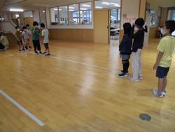 玄関ホールの木製の床に引かれた白い線の上に、マスクを着用した複数の子どもたちが左右に分かれて列になって立っている様子の写真