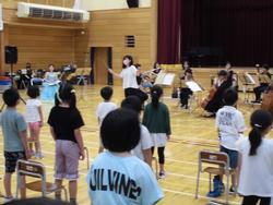 第2学年担任の指揮のもと,オーケストラの演奏に合わせて、椅子の前に立った子どもたちが歌を歌っている様子の写真