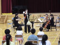 体育館で、バイオリンやチェロ、箒を使って演奏する4人の楽団員と、それらを椅子に座って聞いている子どもたちを捉えた写真