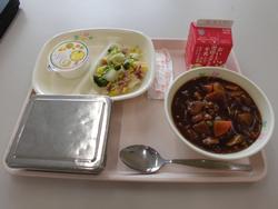 ピンクのトレイの上に、ごはん、ハヤシライス、ブロッコリーとベーコンのソテー、セレクトデザート、牛乳パックが並べられた給食の写真