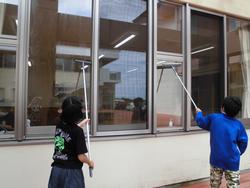 屋外で、2人の子どもたちが柄の長い窓拭き用ワイパーを使い、校舎の窓ガラスを熱心に清掃している写真