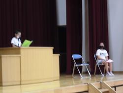 舞台の上で、一人の生徒が椅子に座って待っており、もう一人の生徒が演台に立って原稿を読み上げている写真