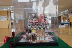 透明なケースに入れられ、横から見た屋根にマカロンやクリームの飾りが並ぶお菓子の家の写真