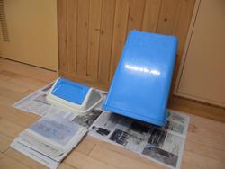新聞紙の上に、青色のごみ箱が逆さまになって壁に立てかけて置かれている写真