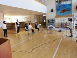 女子児童が前に立って本の紹介するのを、椅子に座り聞いている児童や立って聞いている先生達の朝会の様子の写真