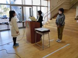 演壇で先生が賞状を読み上げ、向かい合わせに表彰された女の子が真っすぐ立っている写真
