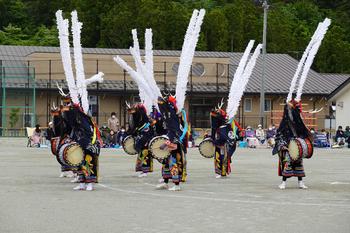 黒い衣装に大きな太鼓をつけ、頭に白く長い飾りを立てた踊り手たちが円を作って踊っている写真