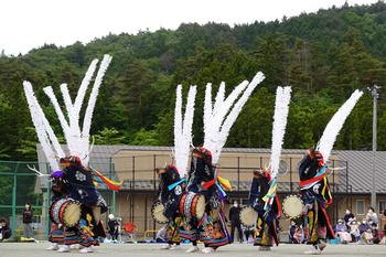 黒い衣装に大きな太鼓をつけ、頭に白く長い飾りを立てた子供達が円を作って踊っている様子を近くから撮影した写真