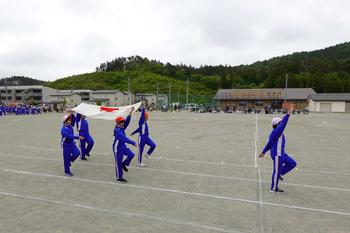 青いジャージを着た児童が手を上げて歩く生徒に続いて4人で国旗を持って入場行進している写真