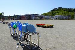 手前に水やりするジョウロが傘立てに並べられ、奥に花の苗が運動場に置かれた写真