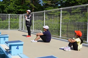 スタート台が設置された屋外プールのプールサイドに、白いキャップと赤いキャップをかぶった小学生たちが並んで座り奥の女の子がマイクを使い発表をしている写真