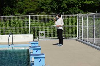 屋外プールのプールサイドで女性教諭がマイクを持ち説明をしている写真