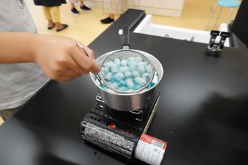 黒い作業台の中央に設置された卓上コンロに置かれた鍋の中で、染色液の中に入れた繭が水色に染まり、児童がザルでかき混ぜている様子の写真