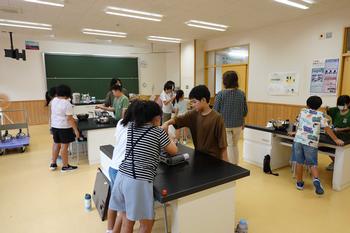黒板や理科の実験器具などが置かれた理科室で、女性講師の指導のもと、グループごとに分かれた複数の児童たちがテーブルを囲んで作業している様子の写真
