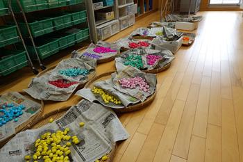 新聞紙が敷かれた竹ざるの上に黄色やピンク色など様々な色が付けられた繭玉を、フローリング床の上に並べて乾かしている様子の写真