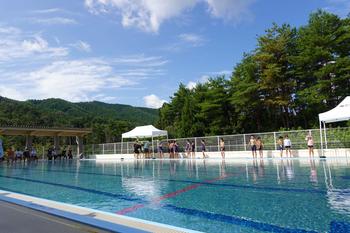 奥に緑豊かな木々や山が見える青空の下、白いテントがいくつか設置されているプールサイドに水着姿の大勢の子児童たちが立ち並んでいる写真