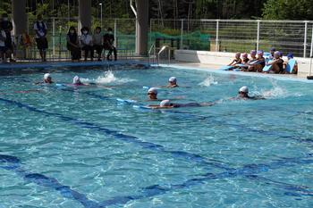 プールサイドで、他の児童たちや先生らしき大人が見守っているなか、白い水泳帽を被った6人の児童たちが水色のビート板を両手で持ち泳いでいる校内水泳記録会の様子の写真