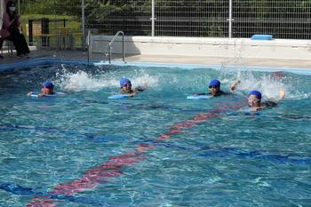 日陰に座って応援している大人が見守る中、青い水泳帽をかぶっている4人の児童たちがビート板を使ってプールで泳いでいる様子の写真