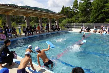 日陰の椅子に座った保護者たちや、プールサイドに座った児童たちが見ている中、順番がきた男子児童がブールに入ったり、水色のビート板を持った児童たちがプールの中でバタ足で泳いでいる様子の写真