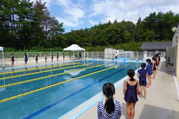 夏のような晴れた空の下、コースロープが張られているプールサイド奥に1人の先生が立ち、水着姿の児童たちが間隔をあけて左右のプールサイドに立ち先生の話を聞いている様子の写真