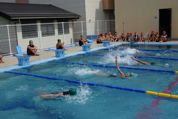 他の子供たちがプールサイドに座って見守っている中、緑色の水泳帽を被った数人の子供たちがプールで泳いでいる写真