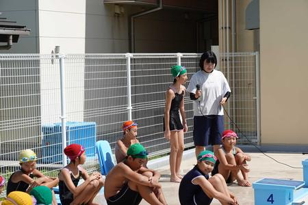 カラフルな色の水泳帽を被った多くの児童たちがプールサイドに座っている中、緑色の水泳帽を被った女子児童が女性教師からマイクを向けられ話をしている写真