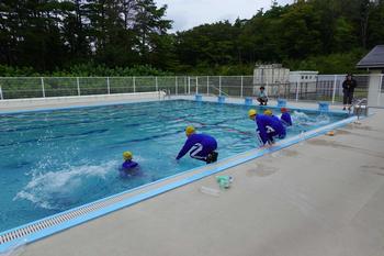 黄色の帽子をかぶりジャージ姿の複数の子供たちが屋外のプールサイドから水しぶきをあげながら入水している様子を写した写真