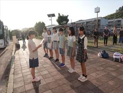 後ろにはバスが停まっており、旅行用のカバンも置かれている広い歩道で、整列した6人の児童が、向かい合って立っているノートの様な物を持ちながら話をしている1人の女の子の話を熱心に聞いている出発式の様子の写真