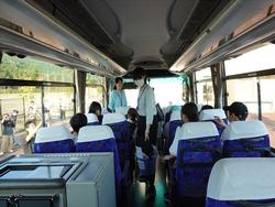 窓の外には多くの大人が手を振って見送っているなか、バスの通路には引率者の女性が立ち、数人の児童たちが座席に座り窓の外に向かって手を振っているバスの車内から撮影された写真