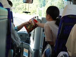 バスの前列の座席に座っているマスクを着用している女の子が、通路を挟んだ座席の人物からスナック菓子の袋を受け取っている様子の写真