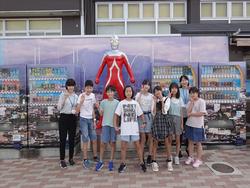 自動販売機が並んでいる間に設置されたウルトラセブンの像の前に引率者の女性と複数の子供たちが整列し一緒に記念撮影をしている写真