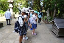 木々に囲まれ、石灯篭が設置された境内で、引率の先生やキャップを被りリュックを背負った子供たちが手を合わせてお参りをしている様子を横から撮影した写真