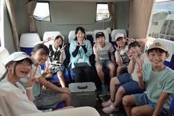 バスの中で、数人の子供たちが引率者らしき女性と一緒に座って、カメラに向かってポーズを取っている写真