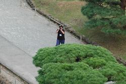 緑に茂った木々に囲まれた通路に立つ黒いTシャツと青いパンツを履いている男性が、カメラを構えて天守閣を撮影している様子の写真