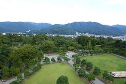 少し曇りがちの空の下、奥に建物が立ち並ぶ市街地とその背景にある山々が見え、手前には芝生の広場や木々が広がる周囲の様子を鶴ヶ城の天守閣から撮影した写真