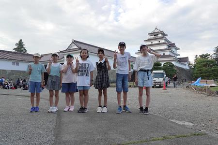 白壁の鶴ヶ城をバックに、横一列に並んだ7人の子供たちがカメラに向かってポーズを取っている写真