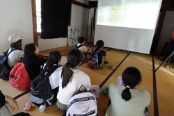 前方のプロジェクターで映像が映し出されたスクリーンを、リュックを背負っている子供たちが和室の床に座って観ている写真
